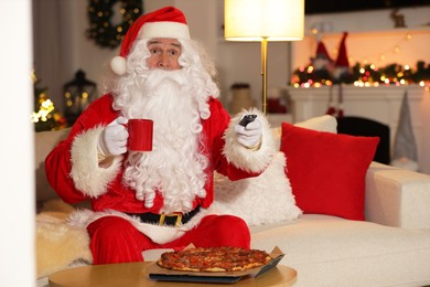 Photo of Merry Christmas. Santa Claus with cup of drink changing TV channels on sofa at home