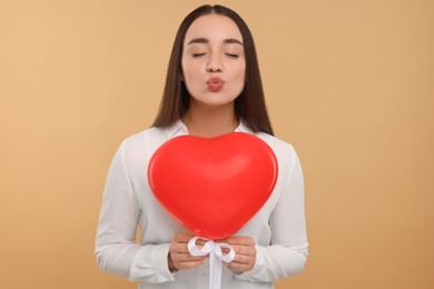 Photo of Young woman holding red heart shaped balloon and blowing kiss into camera on beige background