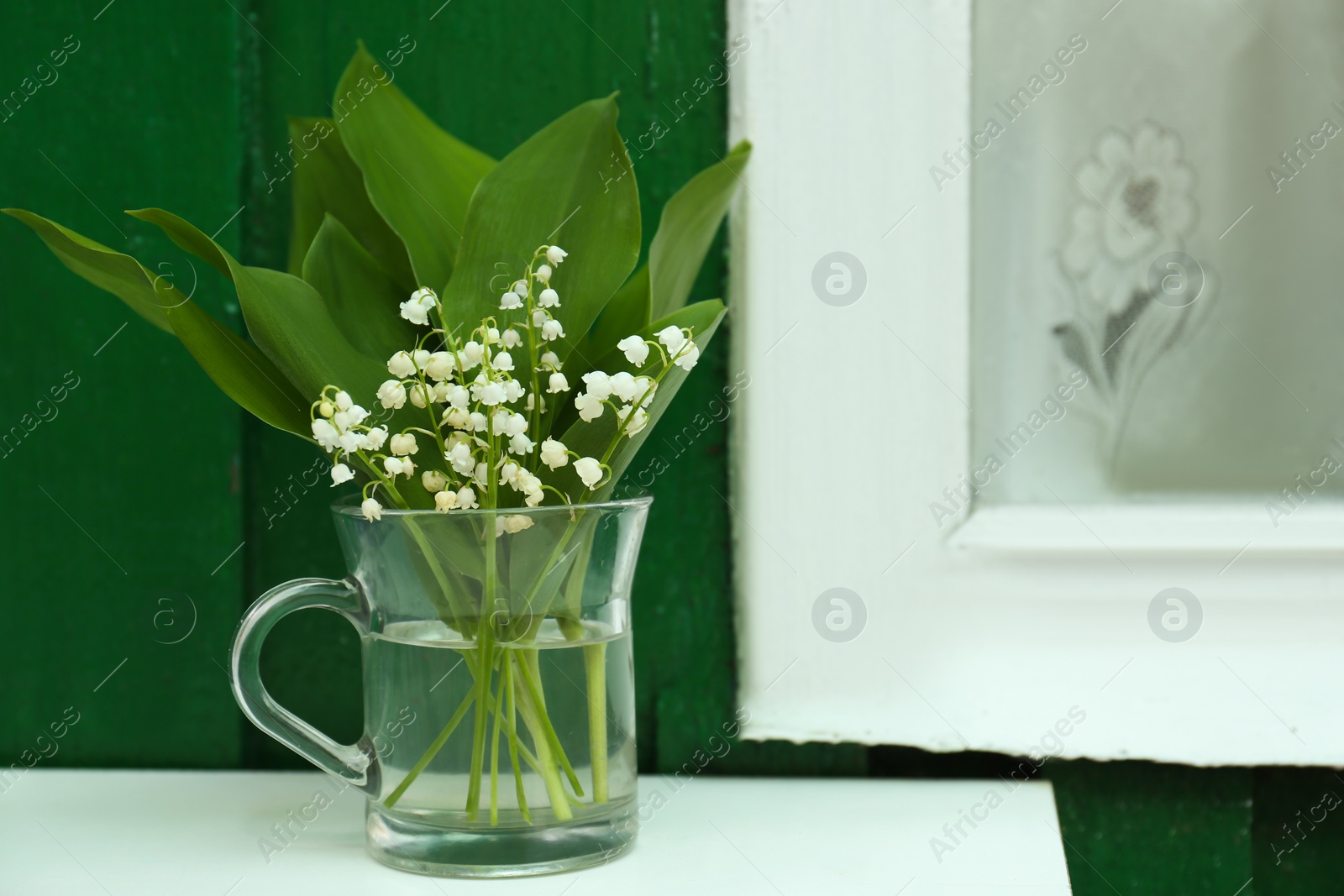 Photo of Beautiful lily of the valley flowers in glass vase on table near green wooden wall outdoors, space for text