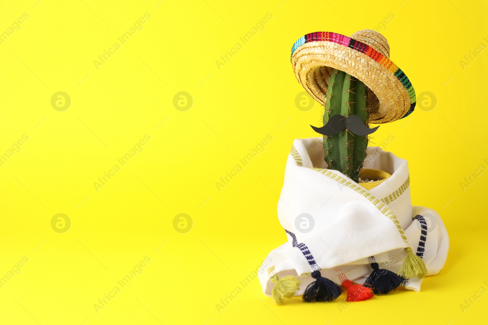 Photo of Cactus with Mexican sombrero hat, fake mustache and poncho on yellow background, space for text