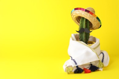 Cactus with Mexican sombrero hat, fake mustache and poncho on yellow background, space for text