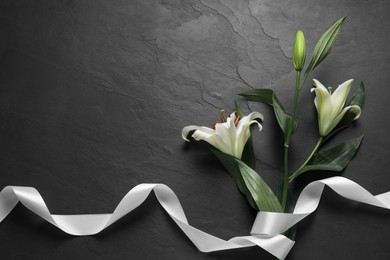 Photo of White lilies and ribbon on black table, flat lay with space for text. Funeral symbols