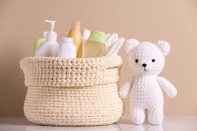 Photo of Knitted basket with baby cosmetic products and toy bear on white table against beige background