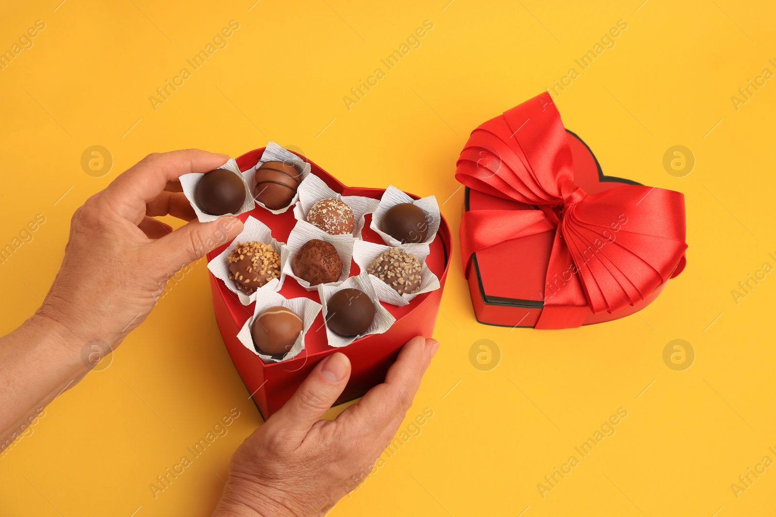 Photo of Woman taking delicious chocolate candy from heart shaped box on yellow background, closeup