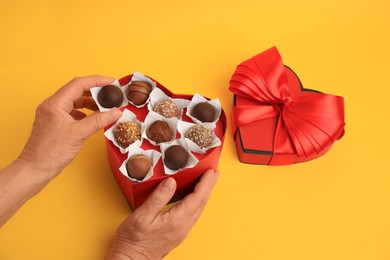 Photo of Woman taking delicious chocolate candy from heart shaped box on yellow background, closeup
