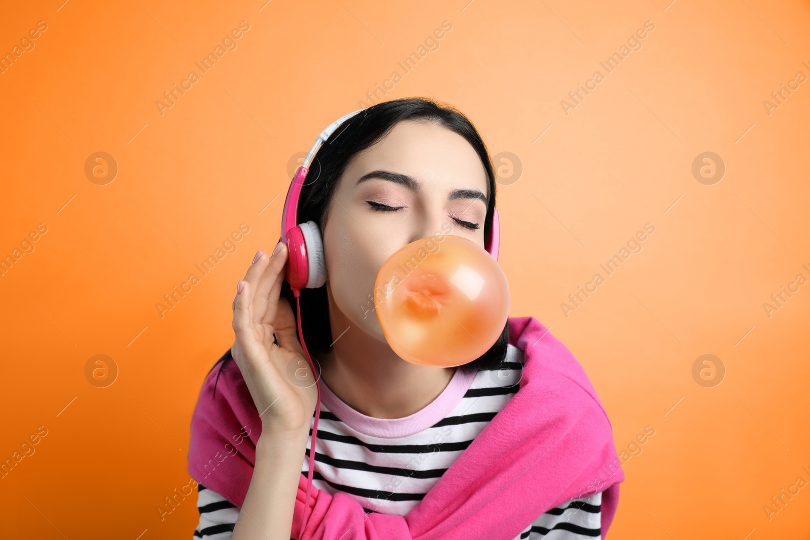 Image of Fashionable young woman with headphones blowing bubblegum on orange background