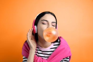 Fashionable young woman with headphones blowing bubblegum on orange background