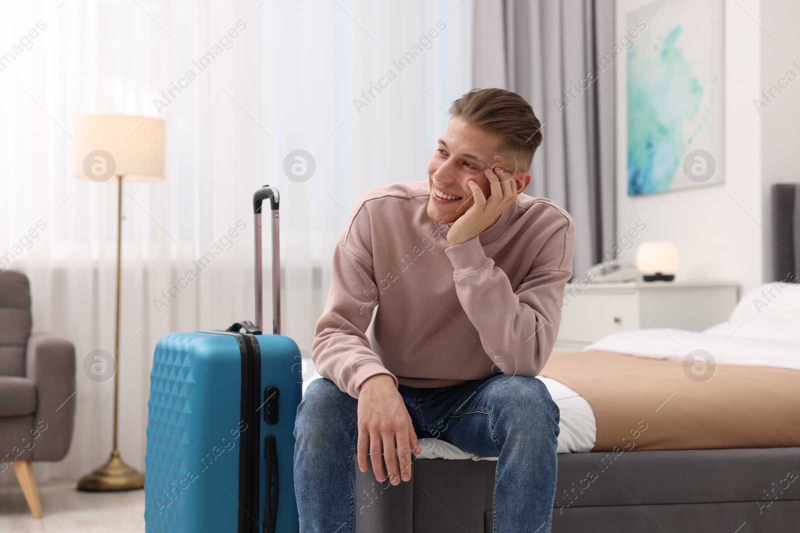 Photo of Smiling guest with suitcase relaxing on bed in stylish hotel room