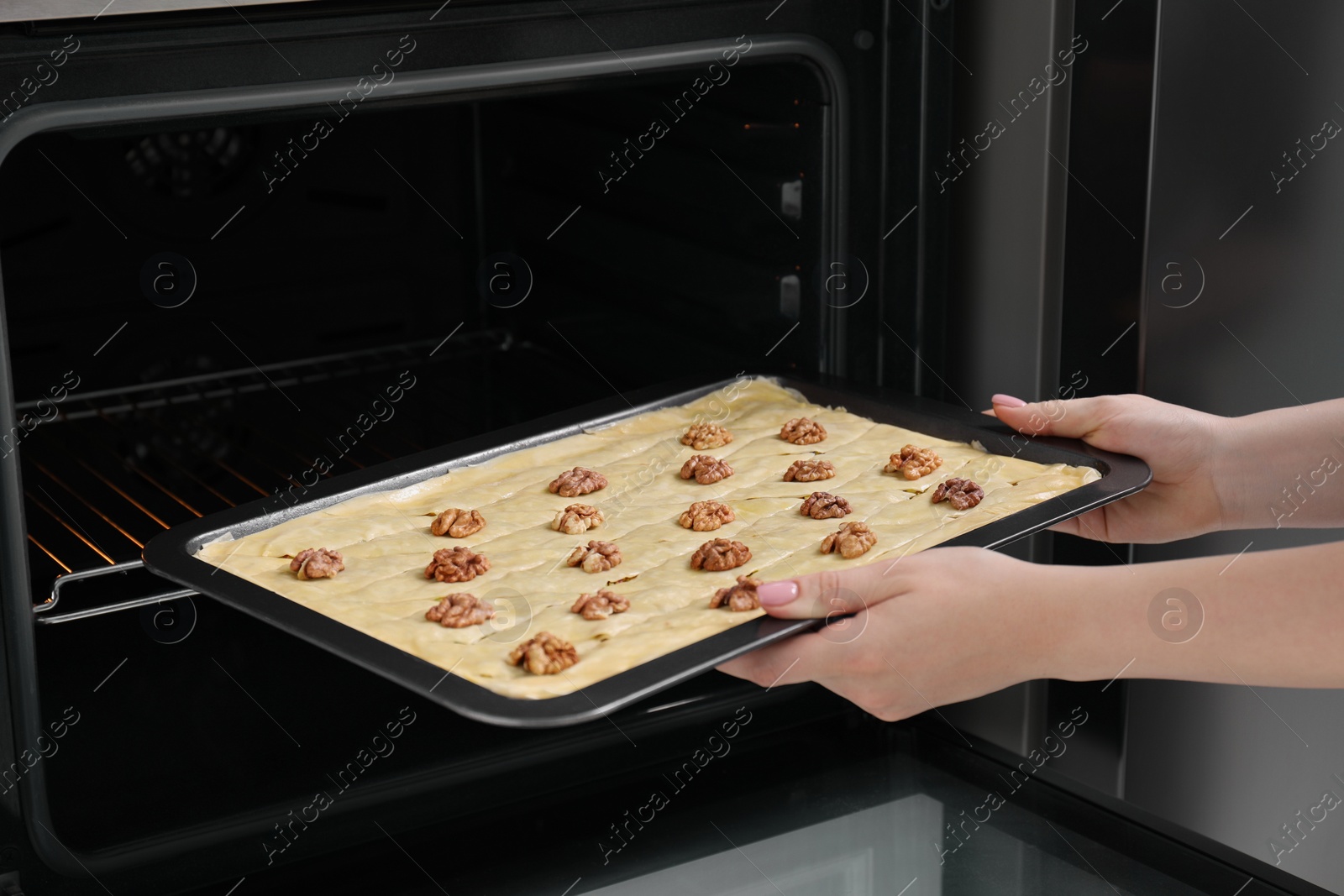 Photo of Woman putting baking pan with baklava into oven, closeup