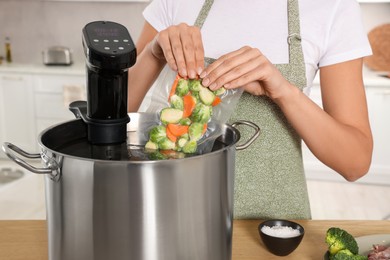 Photo of Woman putting vacuum packed vegetables into pot with sous vide cooker in kitchen, closeup. Thermal immersion circulator