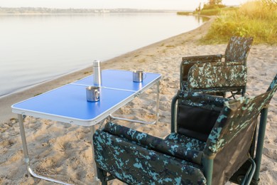 Camouflage fishing chairs and table with metal cups on sandy beach near river