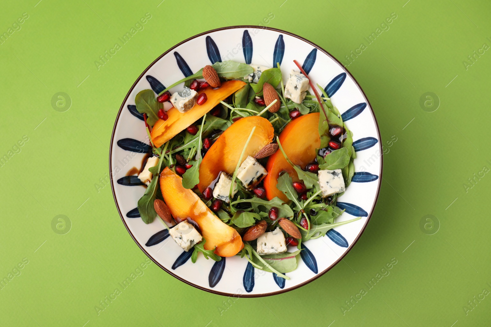 Photo of Tasty salad with persimmon, blue cheese, pomegranate and almonds served on light green background, top view