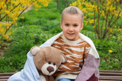 Little girl with teddy bear on wooden bench outdoors