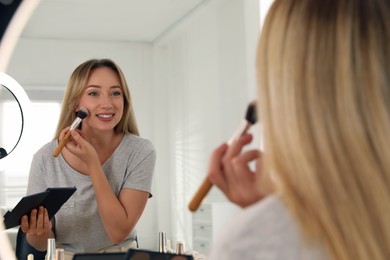 Photo of Beautiful young woman applying highlighter with brush near mirror indoors