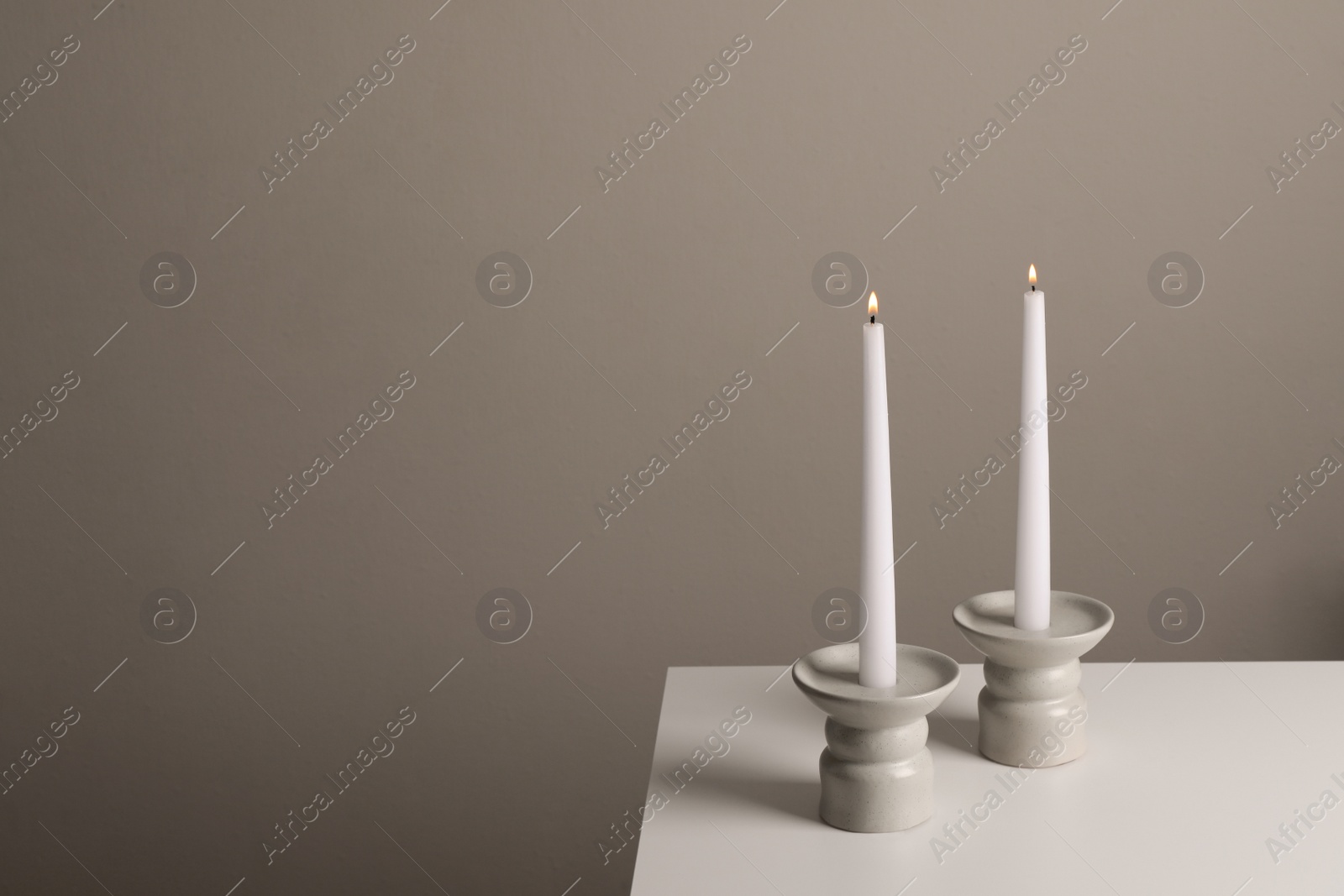 Photo of Holders with burning candles on white table near pale brown wall, space for text