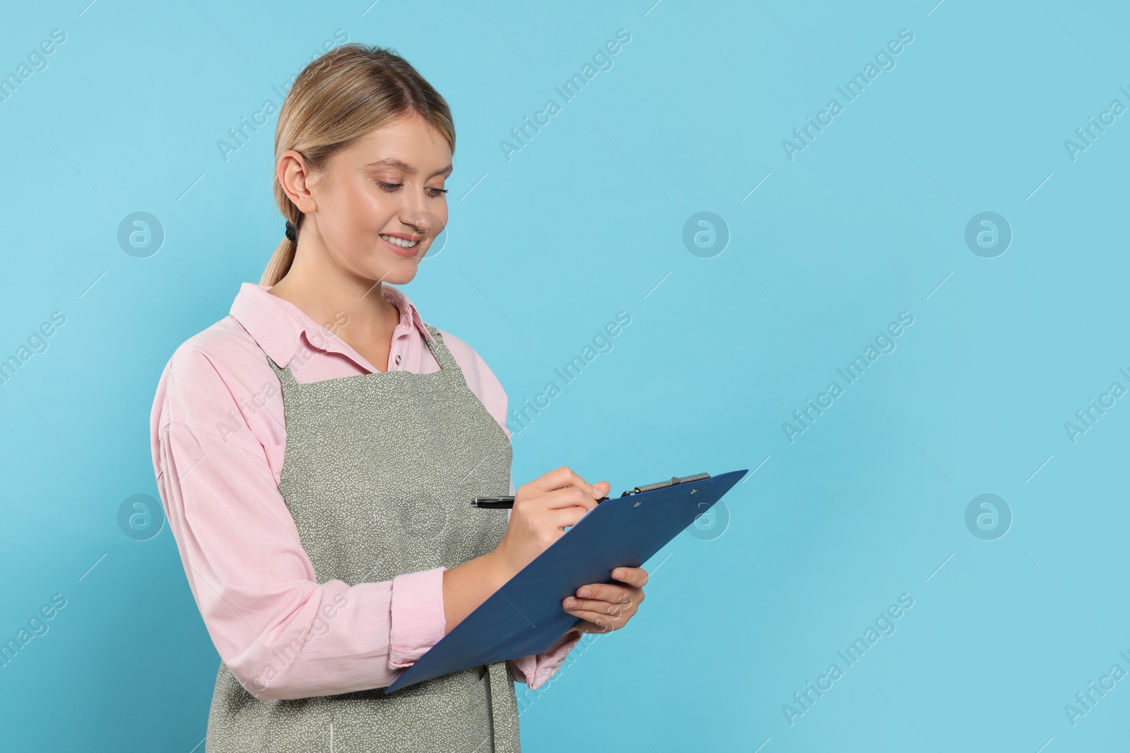 Photo of Beautiful young woman in clean apron with clipboard on light blue background. Space for text