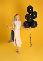 Photo of Happy young woman with balloons and shopping bag on yellow background. Black Friday Sale