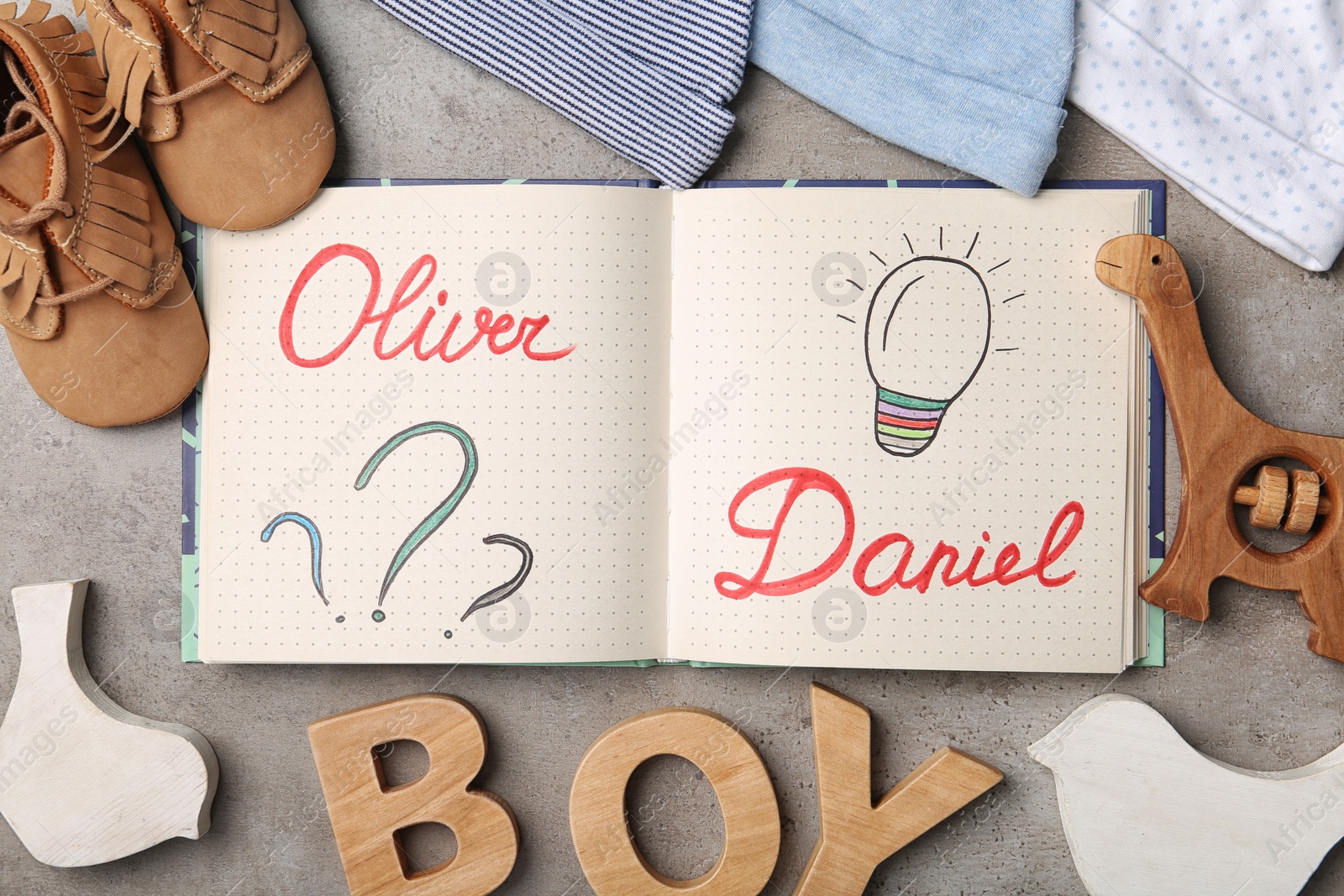 Photo of Notebook with written baby names, child's clothes and wooden letters on grey table, flat lay