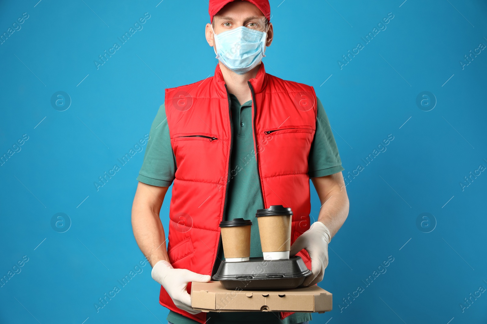 Photo of Courier in protective mask and gloves holding order on blue background. Food delivery service during coronavirus quarantine