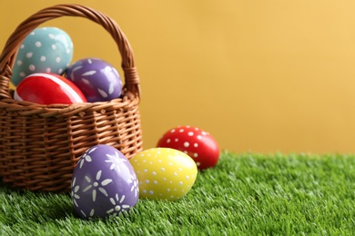 Wicker basket with painted Easter eggs on green grass against color background, space for text