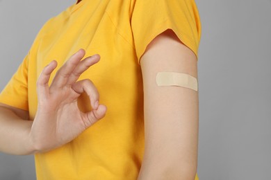 Vaccinated woman with medical plaster on her arm showing okay gesture against grey background, closeup