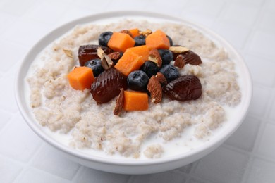Photo of Delicious barley porridge with blueberries, pumpkin, dates and almonds in bowl on white tiled table, closeup