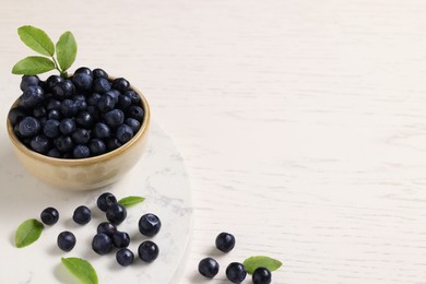 Tasty fresh bilberries with green leaves on white wooden table, space for text