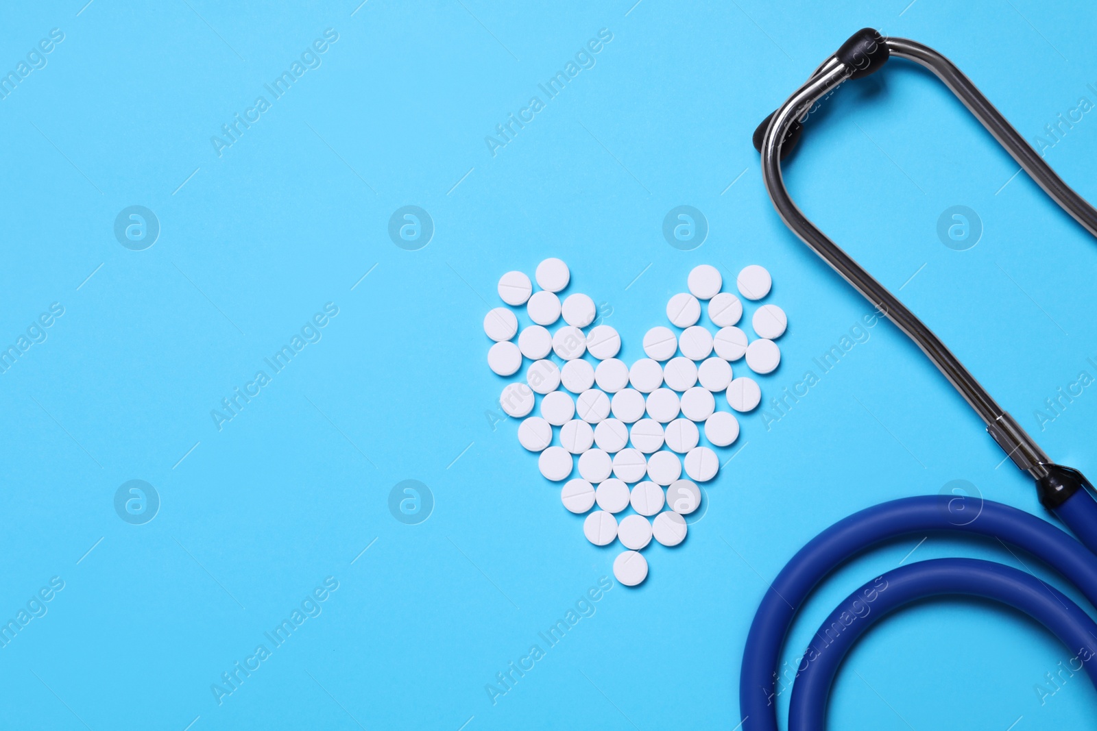 Photo of Heart shape made of pills near stethoscope on light blue background, flat lay. Space for text
