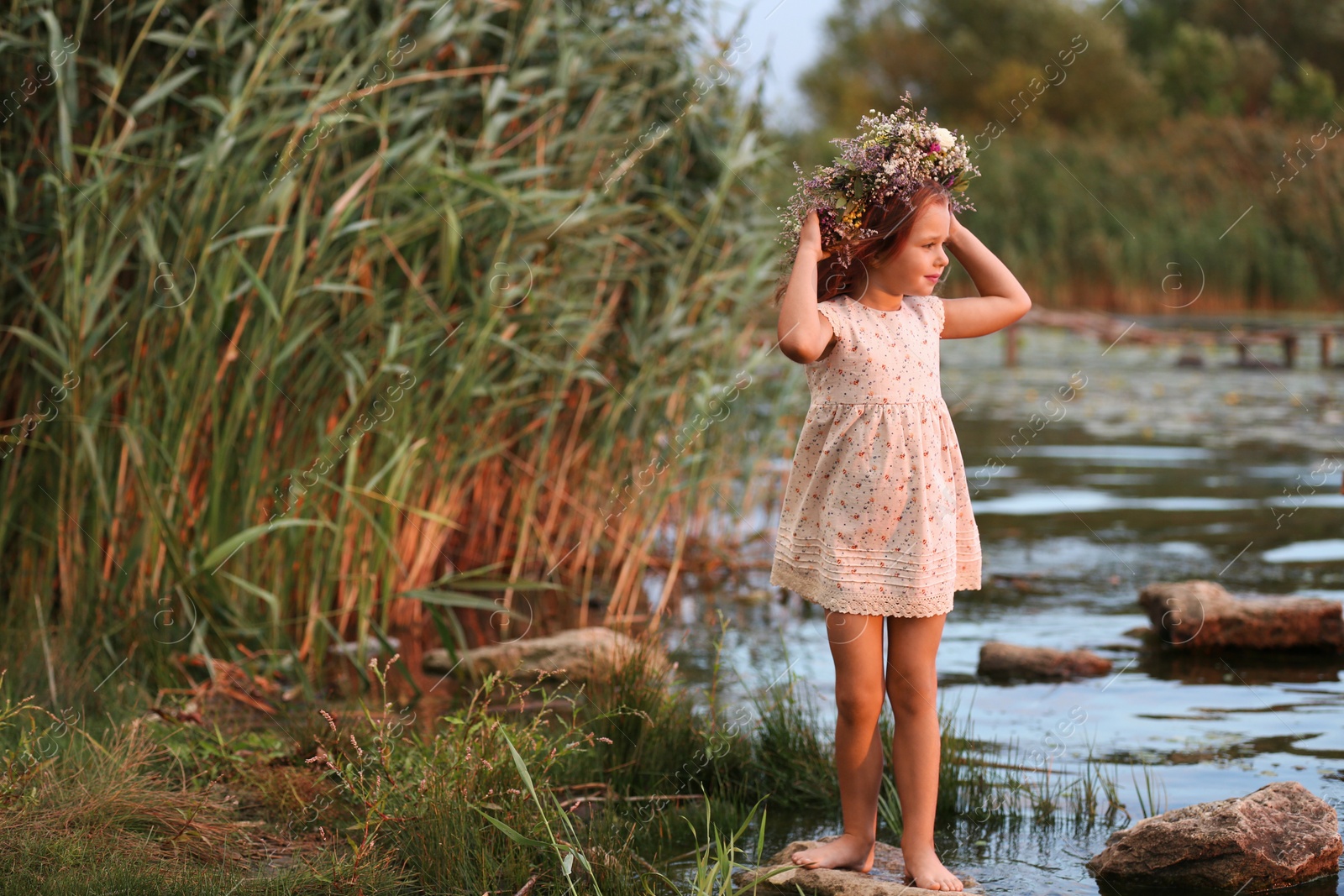 Photo of Cute little girl wearing wreath made of beautiful flowers near river