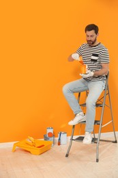 Photo of Designer with painting equipment sitting on folding ladder near freshly painted orange wall indoors, space for text