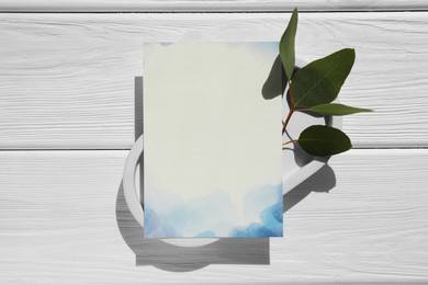 Photo of Blank invitation card and green leaves on white wooden table, top view. Space for text