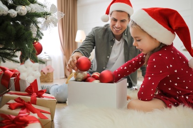 Father with his cute daughter in Santa hats decorating Christmas tree together at home