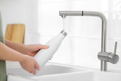 Photo of Woman pouring fresh water from tap into thermo bottle in kitchen, closeup