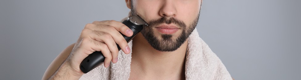 Handsome young man shaving with electric trimmer on grey background, closeup. Banner design