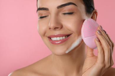 Photo of Young woman using facial cleansing brush on pink background, closeup. Washing accessory