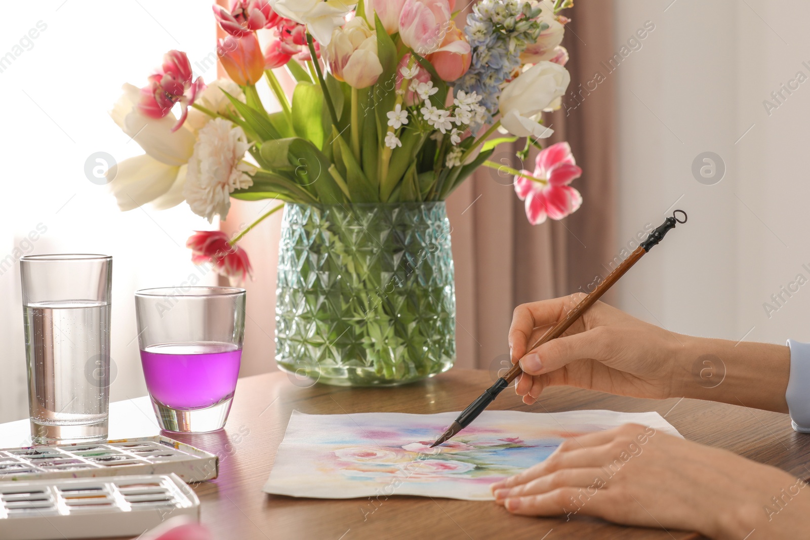 Photo of Woman painting flowers with watercolor at wooden table indoors, closeup. Creative artwork