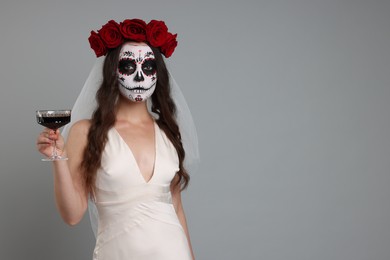 Photo of Young woman in scary bride costume with sugar skull makeup, flower crown and glass of wine on light grey background, space for text. Halloween celebration