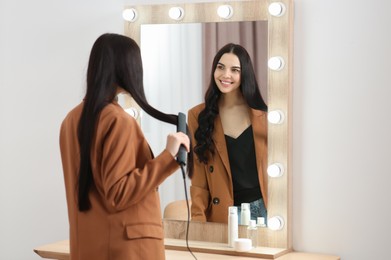 Photo of Beautiful happy woman using hair iron near mirror in room