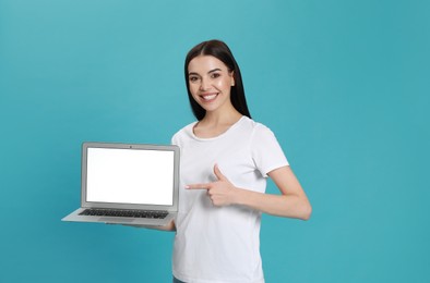 Young woman pointing at modern laptop with blank screen on light blue background