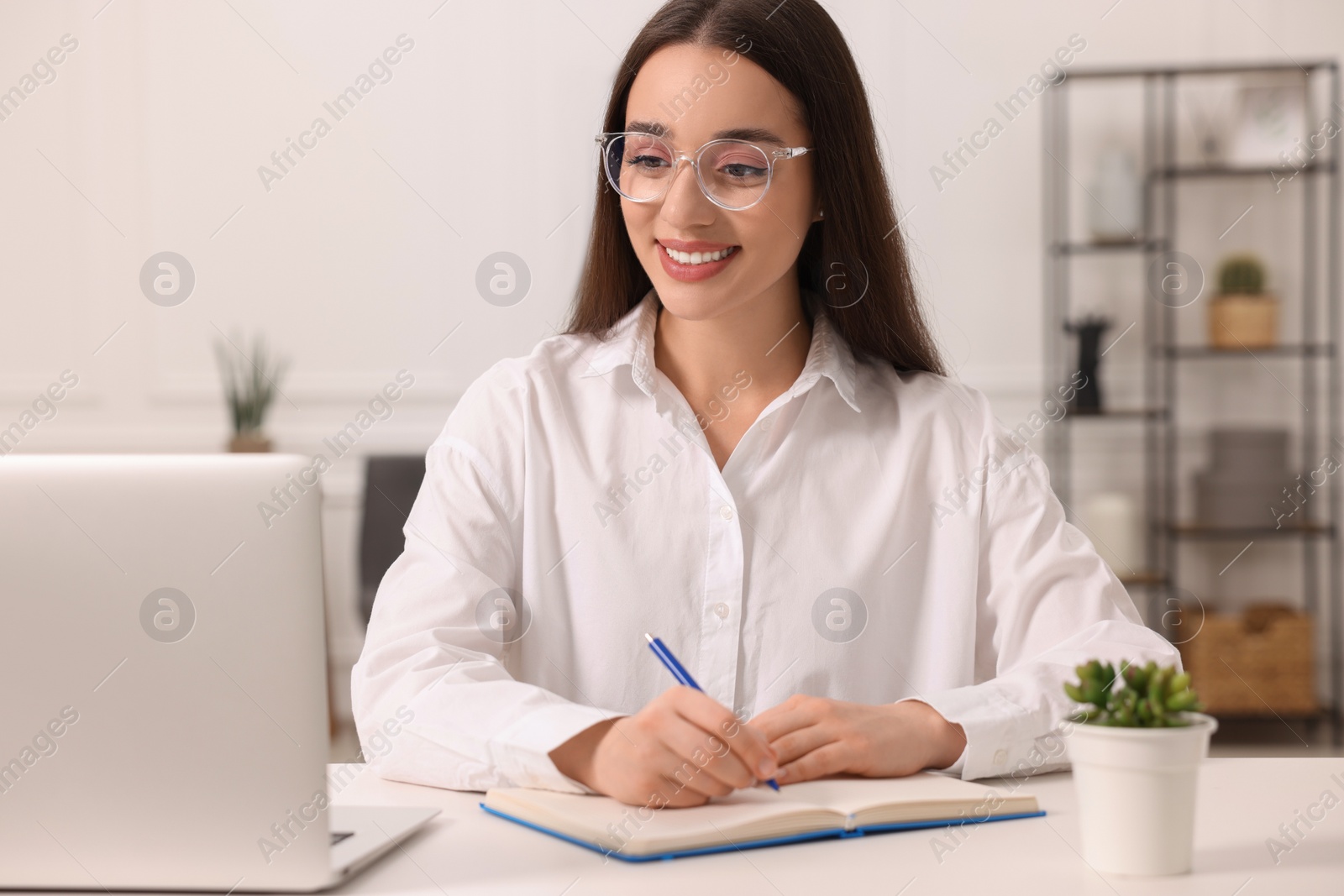 Photo of Young woman writing in notebook at white table indoors