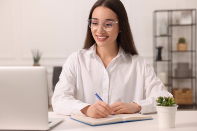 Young woman writing in notebook at white table indoors