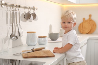 Photo of Curious little boy near kitchen counter with cutting board and sharp knife
