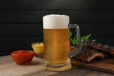 Photo of Mug with beer, delicious grilled ribs and sauces on wooden table