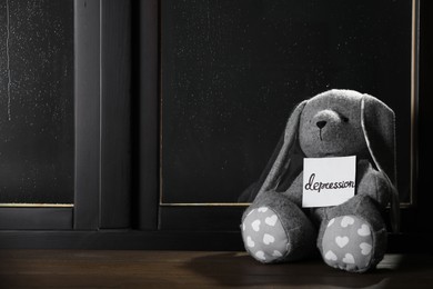 Photo of Toy bunny and card with word Depression near window on rainy night, space for text