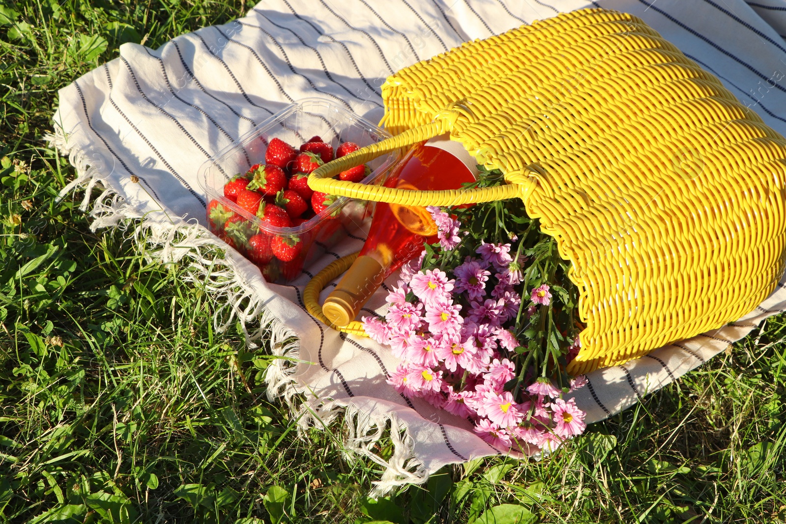 Photo of Yellow wicker bag with beautiful flowers, bottle of wine and strawberries on picnic blanket outdoors