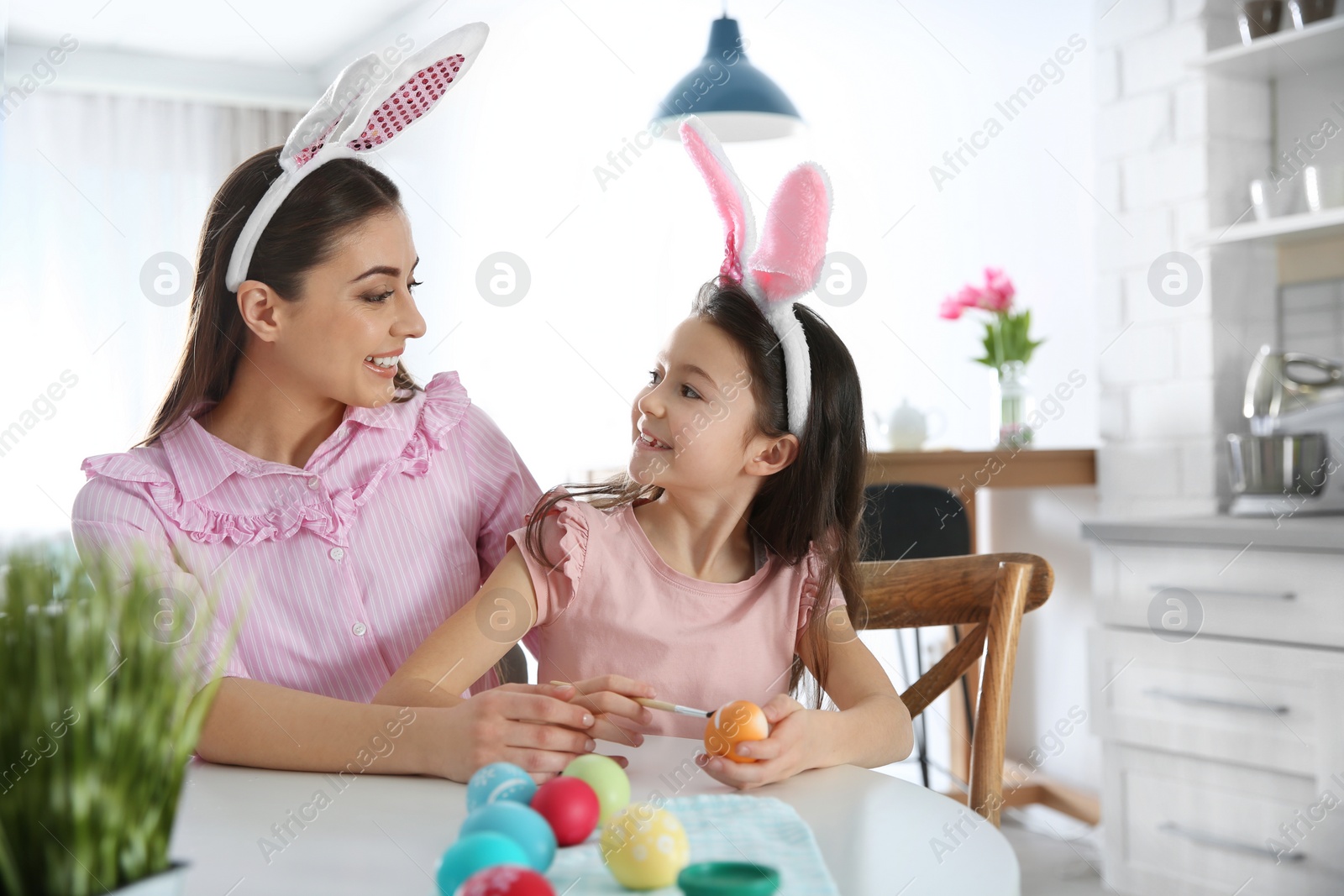 Photo of Mother and daughter with bunny ears headbands painting Easter eggs in kitchen, space for text