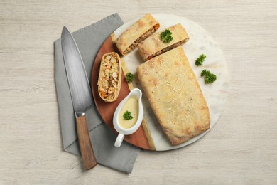 Photo of Cut tasty strudel with chicken, vegetables, sauce and knife on light wooden table, flat lay
