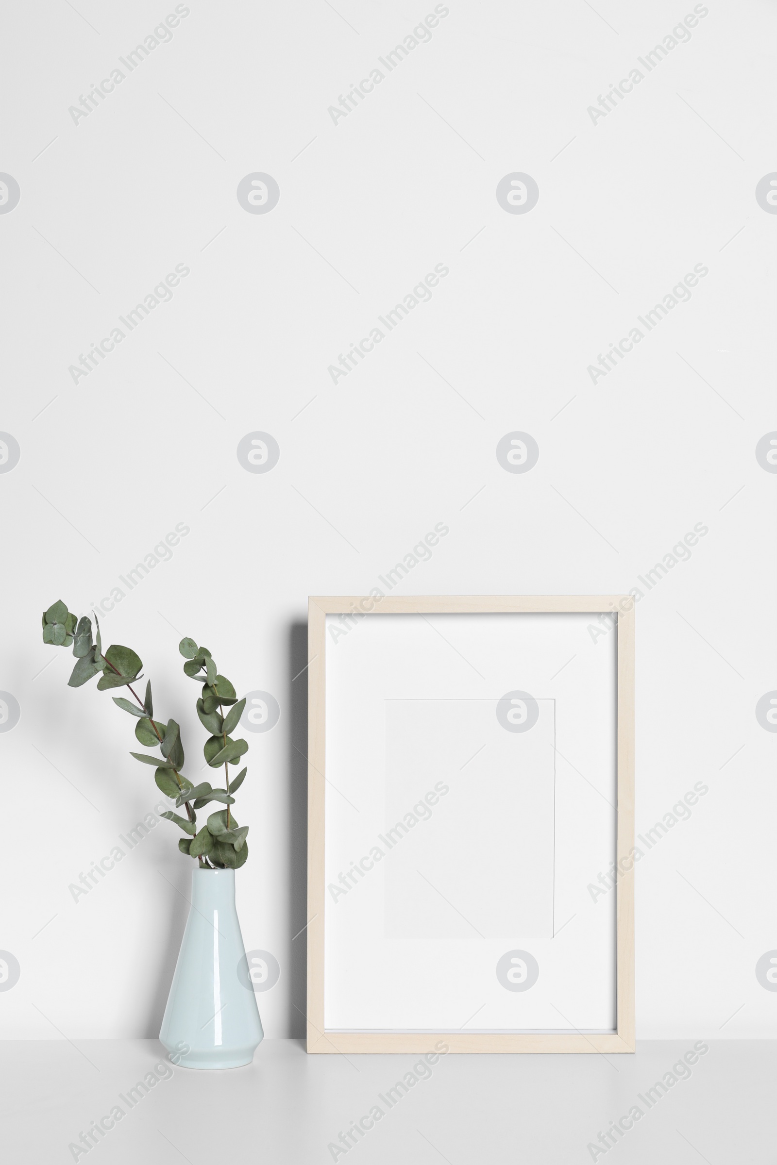 Photo of Empty photo frame and vase with decorative eucalyptus leaves on white table. Mockup for design