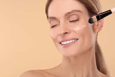 Photo of Woman applying foundation on face with brush against beige background. Space for text
