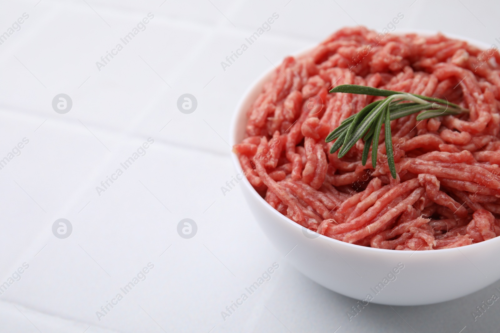 Photo of Fresh raw ground meat and rosemary in bowl on white tiled table, closeup. Space for text